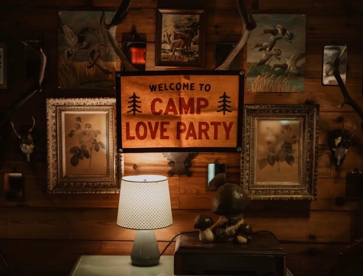Rustic wooden wall with framed artwork, antlers, a lamp, and a sign reading "Welcome to Camp Love Party.