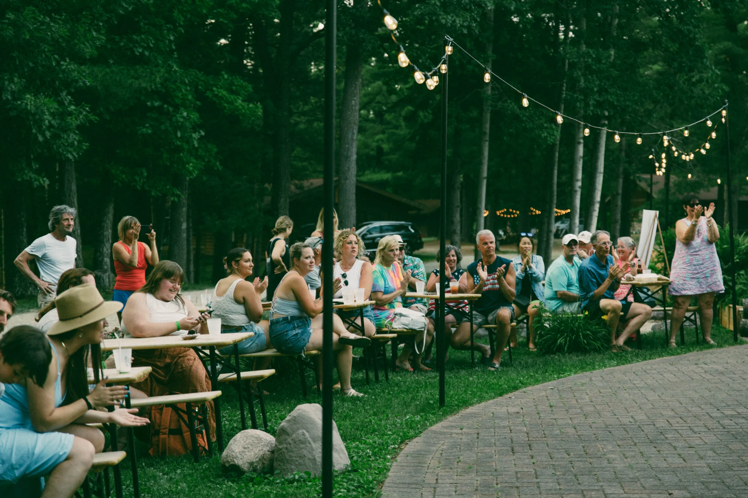 Outdoor gathering of diverse adults seated at picnic tables under string lights, clapping and socializing.