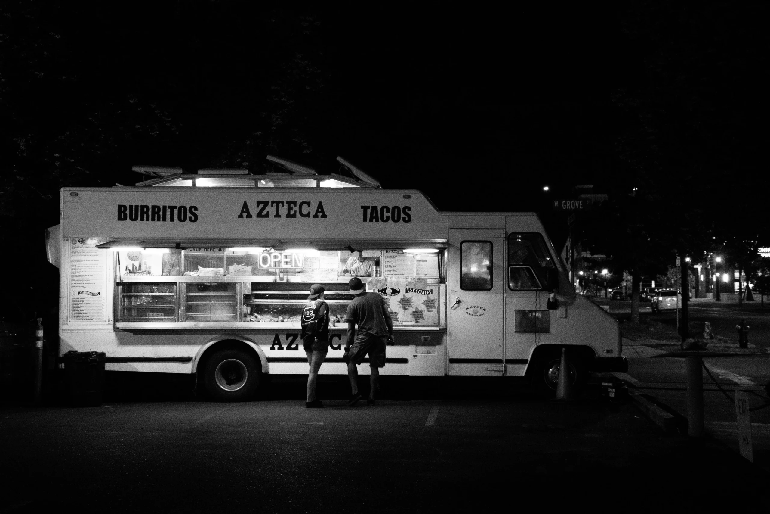 Black and white image of an illuminated Azteca burritos and tacos food truck at night with two customers.
